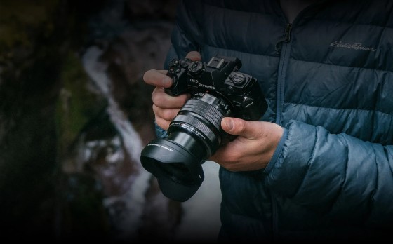 A person holding a camera equipped with the OM System M.Zuiko Digital ED 8-25mm f4.0 PRO for Micro Four Thirds Mount. They are outdoors and there is a waterfall behind them.